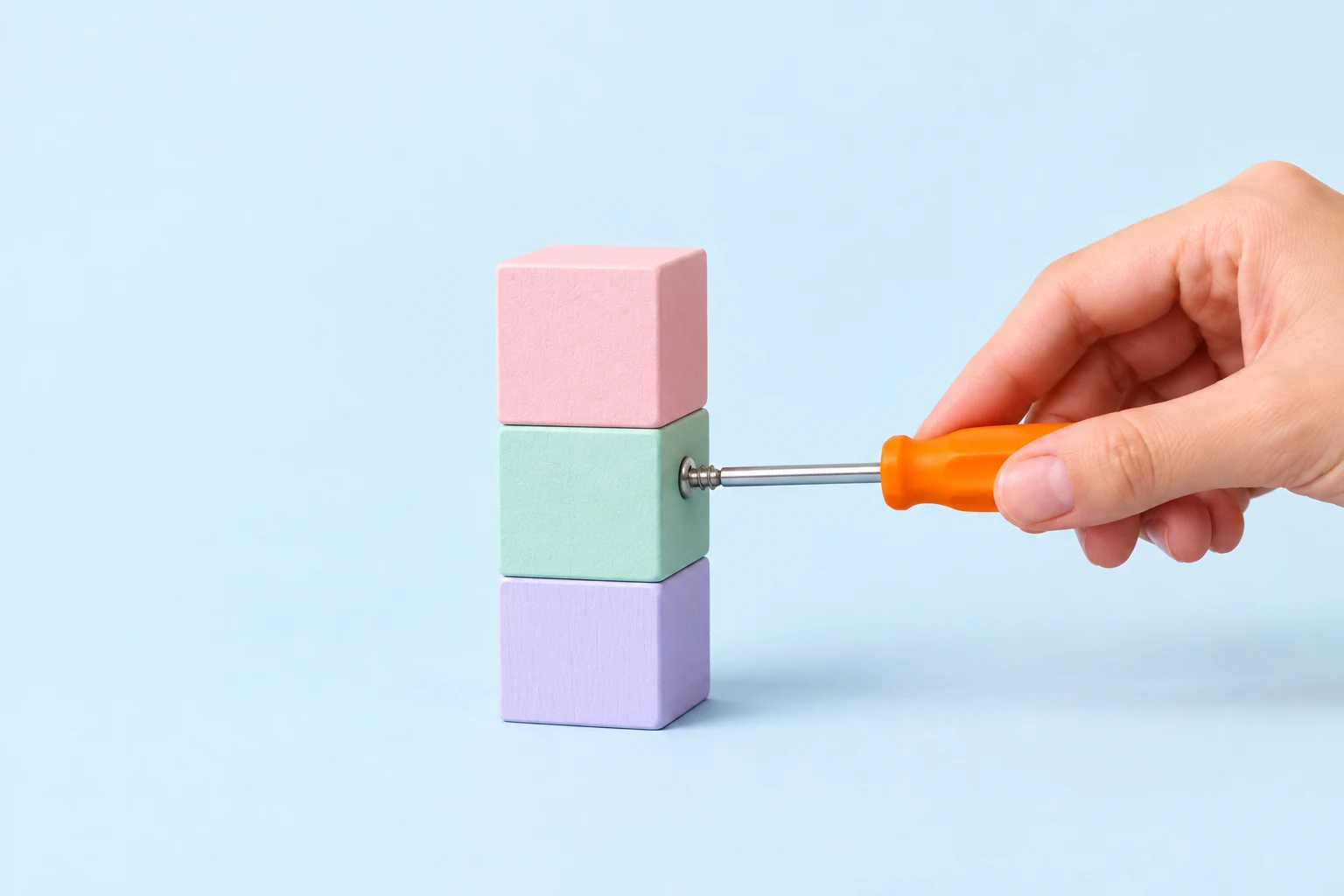 Une main resserre une vis avec un petit tournevis à manche orange dans une pile de blocs en bois pastel