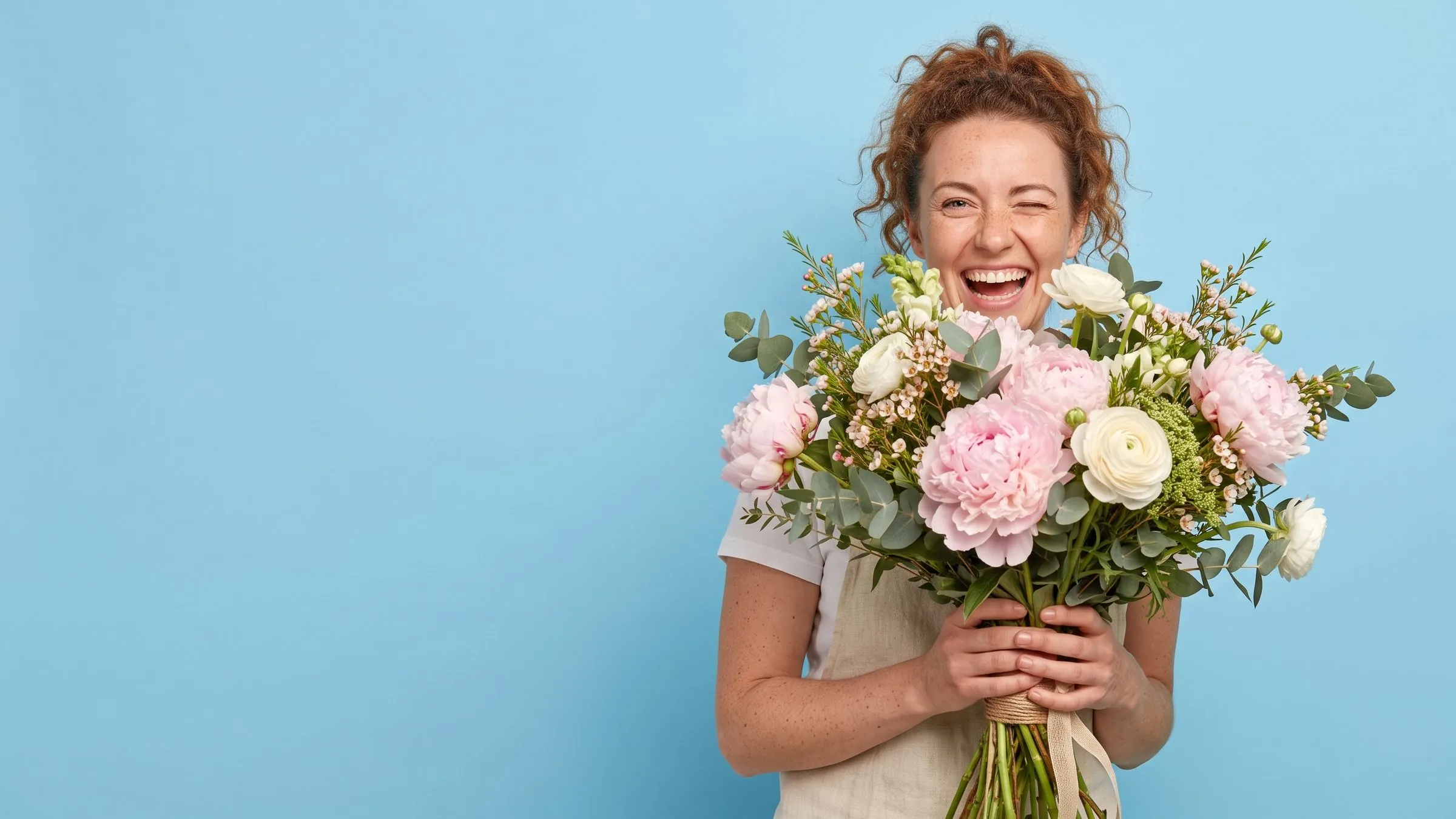 Une fleuriste éclate de rire derrière un grand bouquet de pivoines, sur fond bleu ciel.