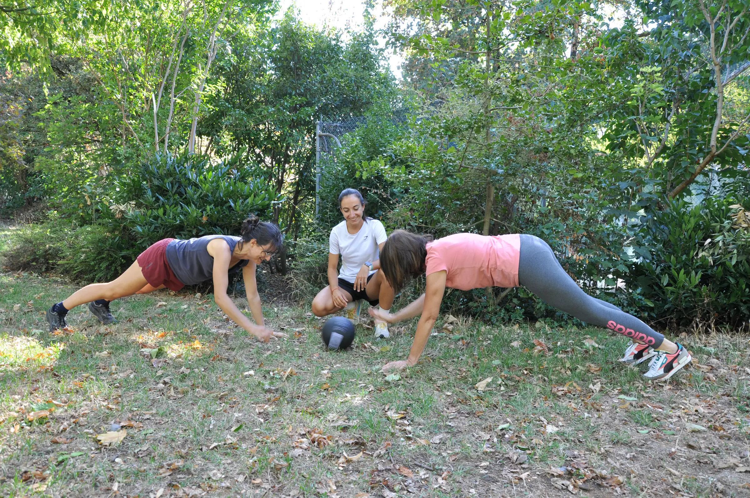 Séance en extérieur sur l’herbe : deux clientes en planche autour d’un médecine-ball, Clémentine accroupie entre elles